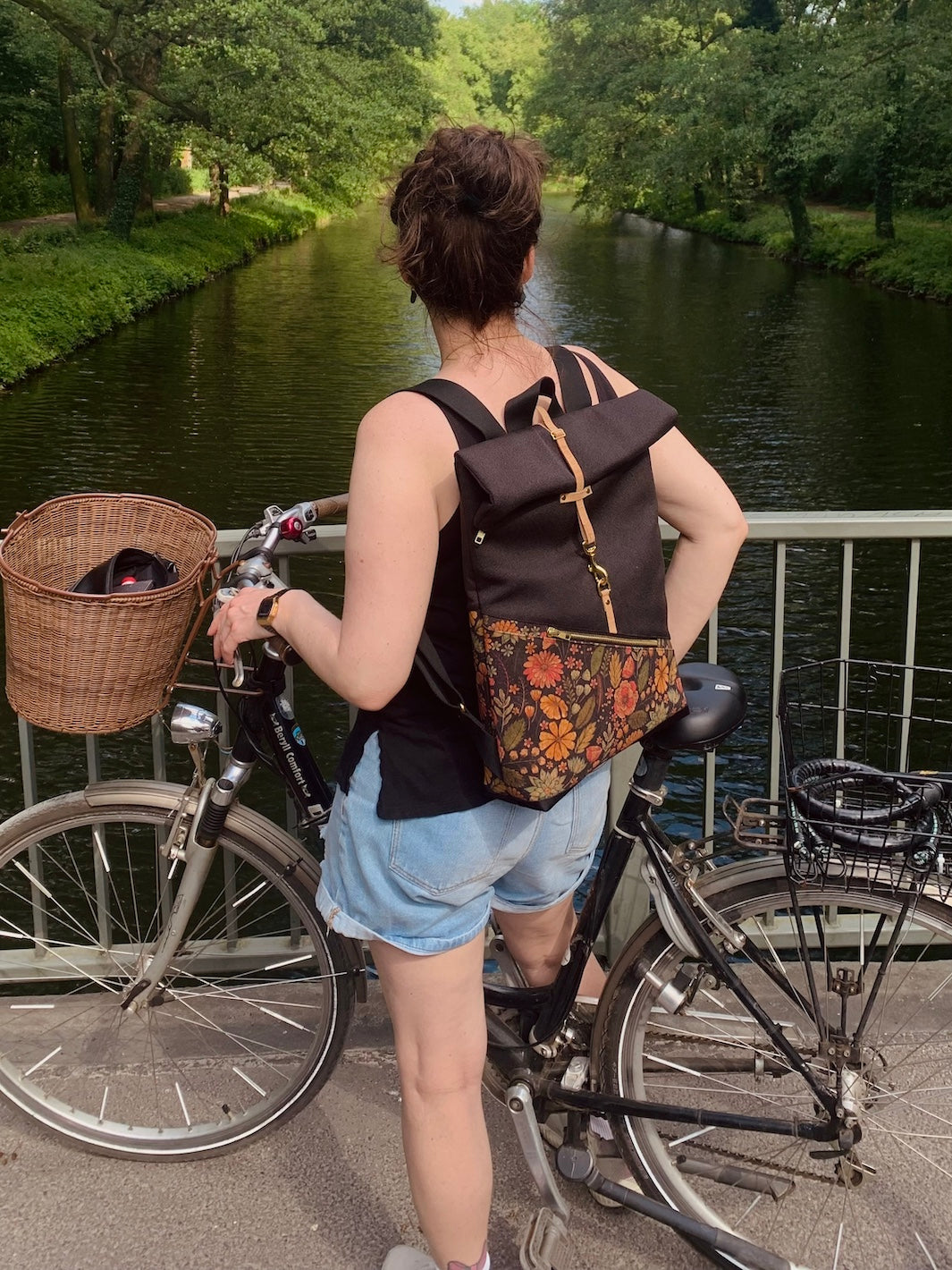 Tragefoto im Sommer: Person mit kleinem Rolltop Rucksack auf dem Rücken auf Brücke mit Fahrrad – urbaner Lifestyle am Fluss.