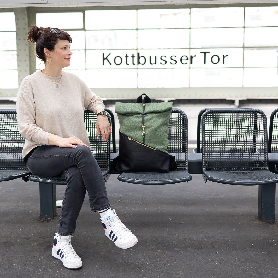 Person sitzt auf der Bank am Kottbuser Tor in Berlin, Rolltop Rucksack neben ihr, mit Blick auf den urbanen Hintergrund.
